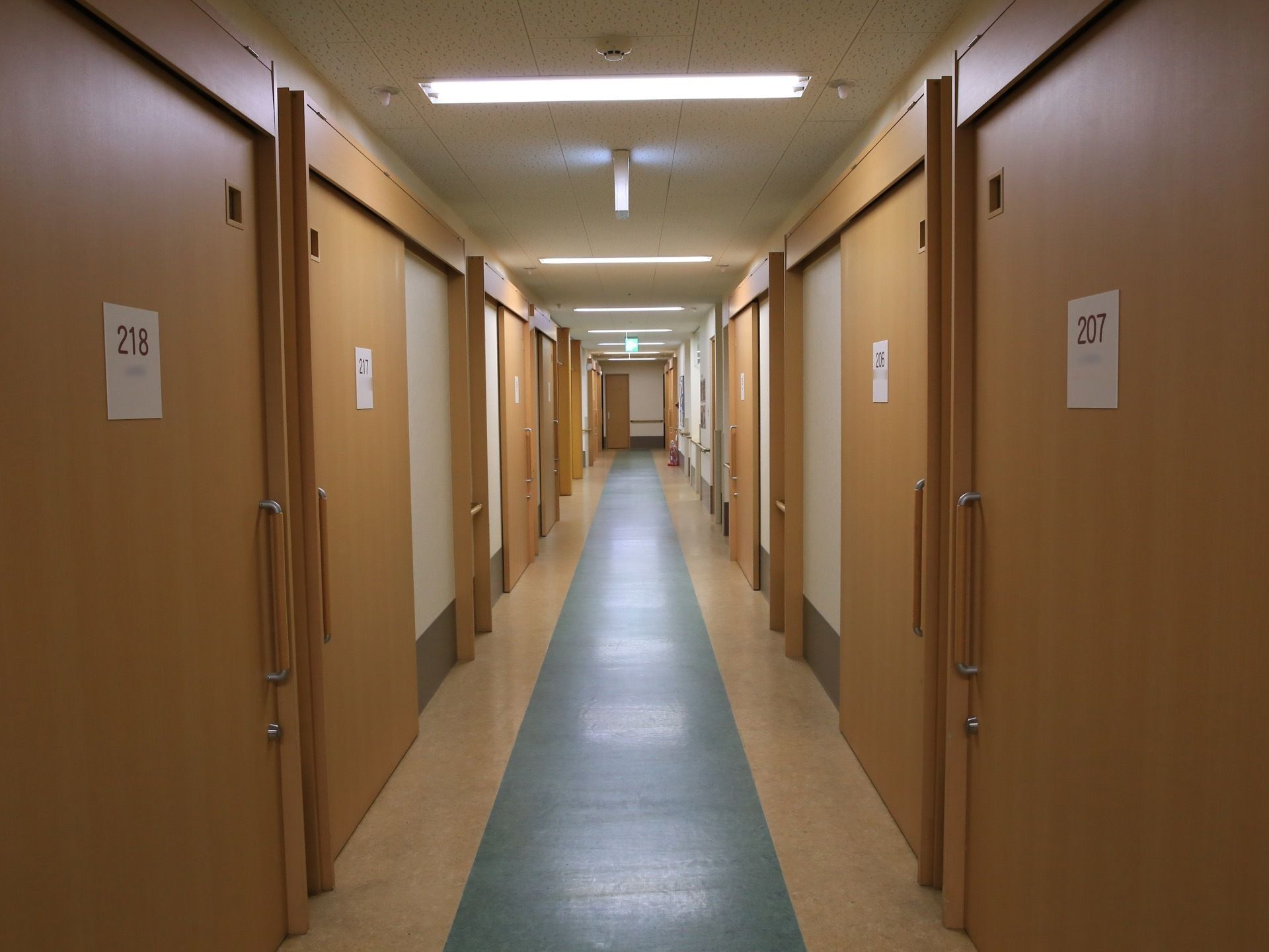 通路の両側に大きな木製の引き戸が並んでおり、扉には部屋番号のプレートが設置されている。天井には、蛍光灯が横向きに連続している。