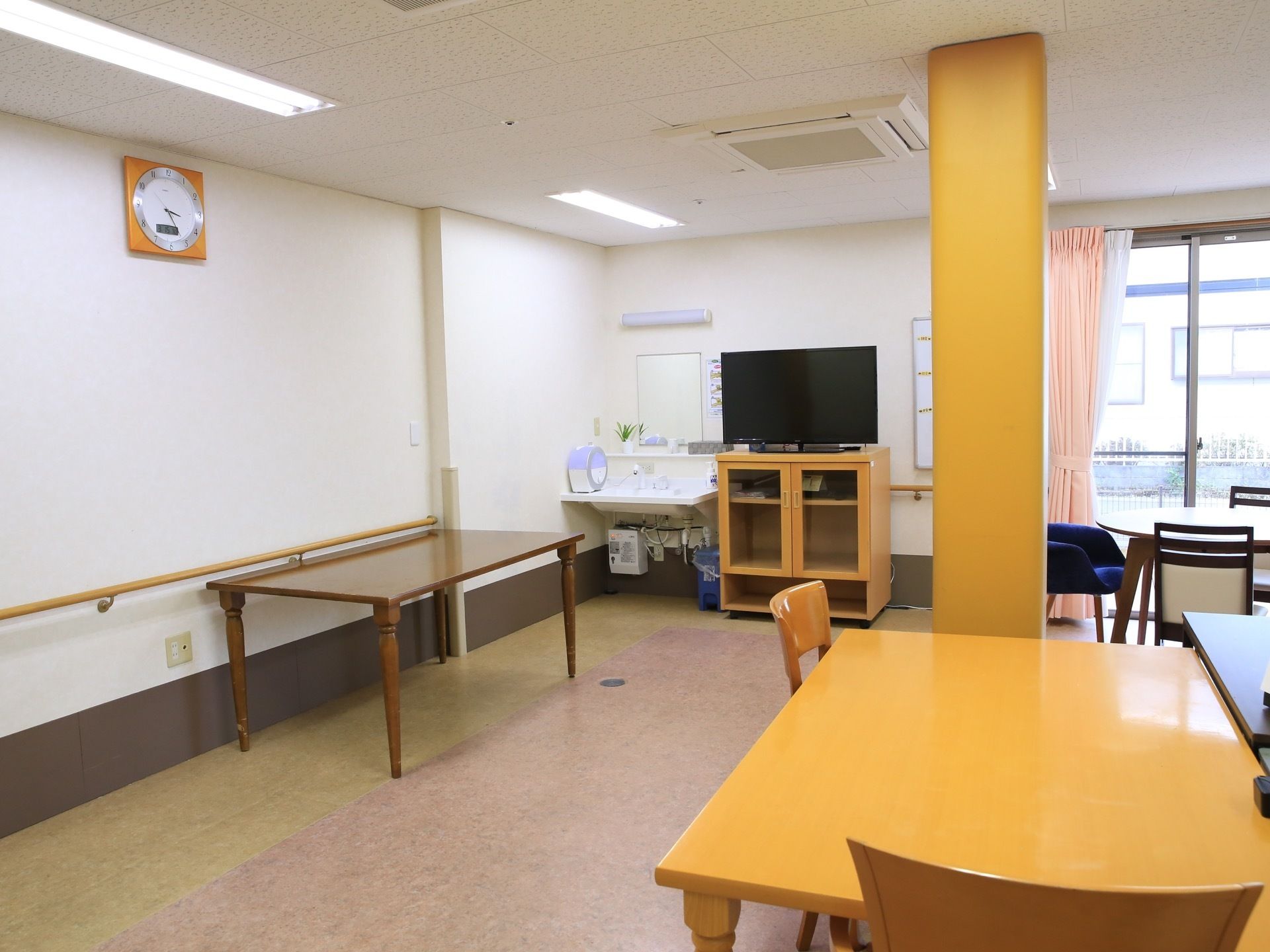 壁掛け時計がある広いフロアにテーブルや椅子が置かれている。左奥に洗面台と鏡があり、その隣のテレビ台にテレビが設置されている。