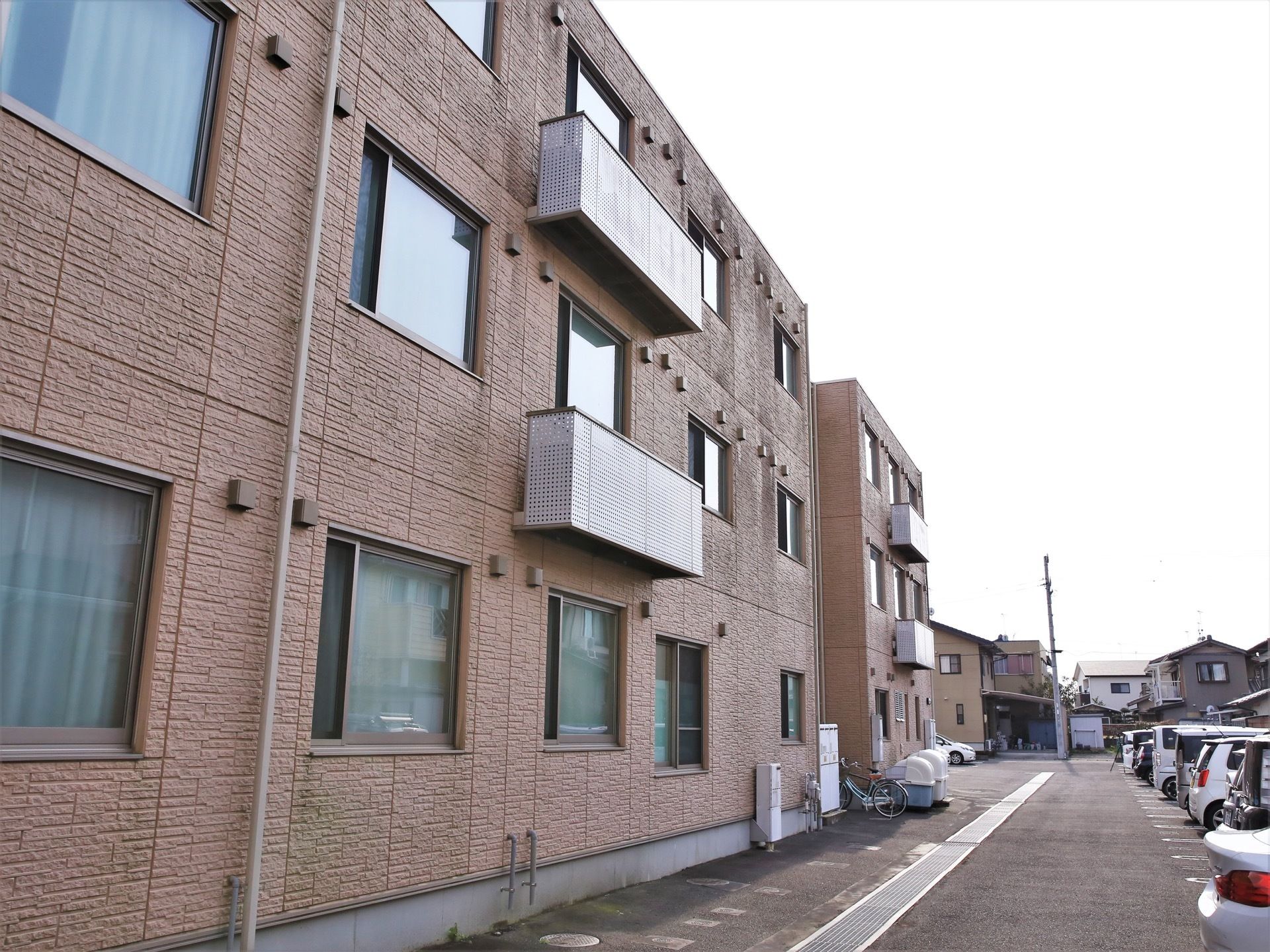 閑静な住宅街の一角に、鉄骨造りの３階建ての施設が建てられている。建物の周囲の路面はアスファルト舗装されており、多くの自動車が停められている。