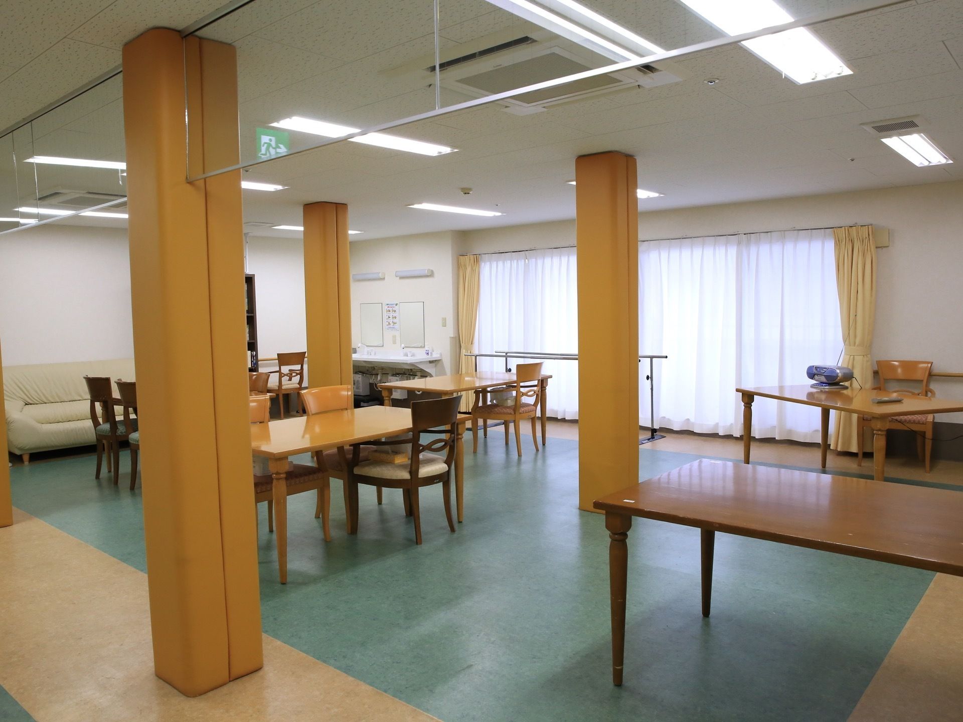 窓のある広間に椅子やテーブルが並べられている。２つ並んだ洗面台、歩行訓練器具、ラジオカセットレコーダーなども見える。