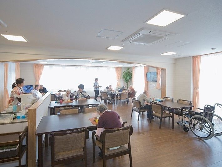 4人掛けのダイニングテーブルが並び、キッチンも設置された明るい雰囲気の食堂。スタッフが所々で介助に当たっている。