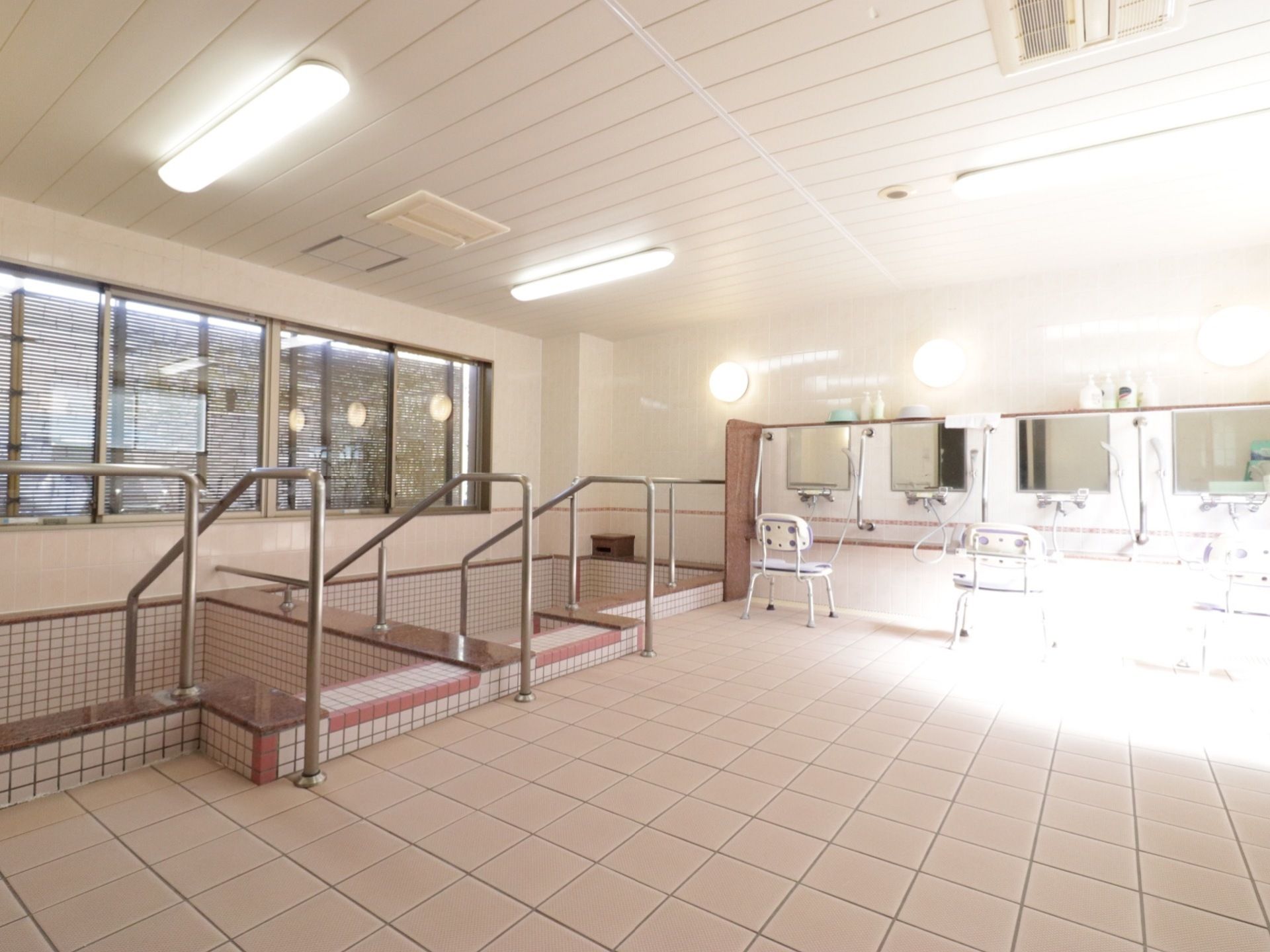 クリームベースの落ち着いた配色の大浴室。手すり付きの大きな浴槽が２つ付いている。各シャワーコーナーにはシャワーチェアが備わっている。