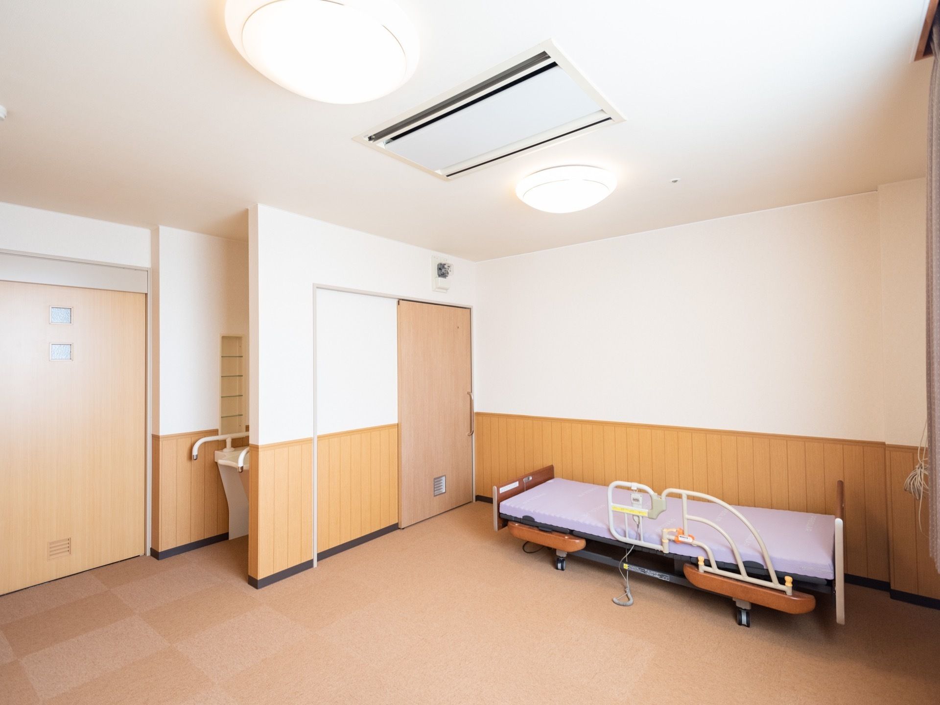 ご入居前の居室である。白い壁に腰高の木目材を採用した壁面となっている。壁際に介護用ベッドが設置されている。