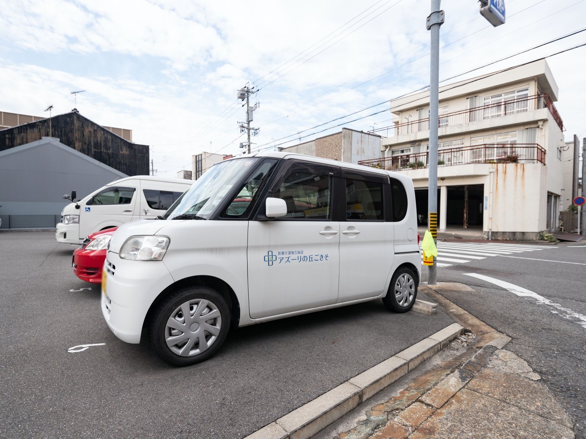 施設の敷地内には、広い駐車場が確保されている。一番端っこには白い社有車があり、隣には赤い車が停められている。