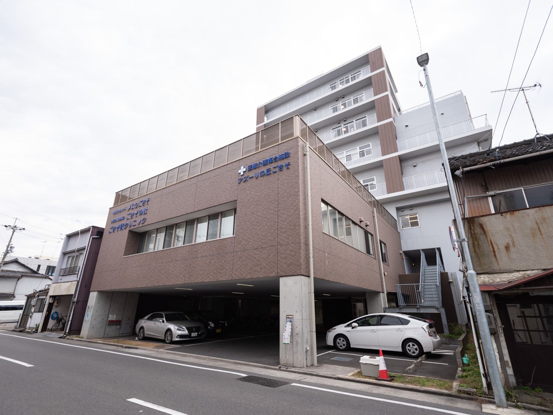 施設は、茶色い外壁で統一された建物となっている。一階部分は駐車スペースになっており、車が３台停められている。