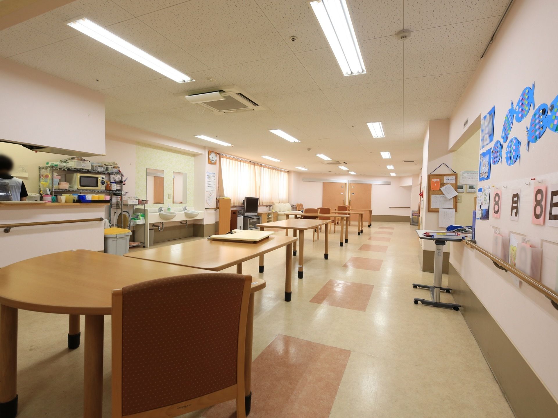 共同生活室は広く、たくさんのテーブルや椅子がある。キッチンが併設されており、スタッフが作業している。