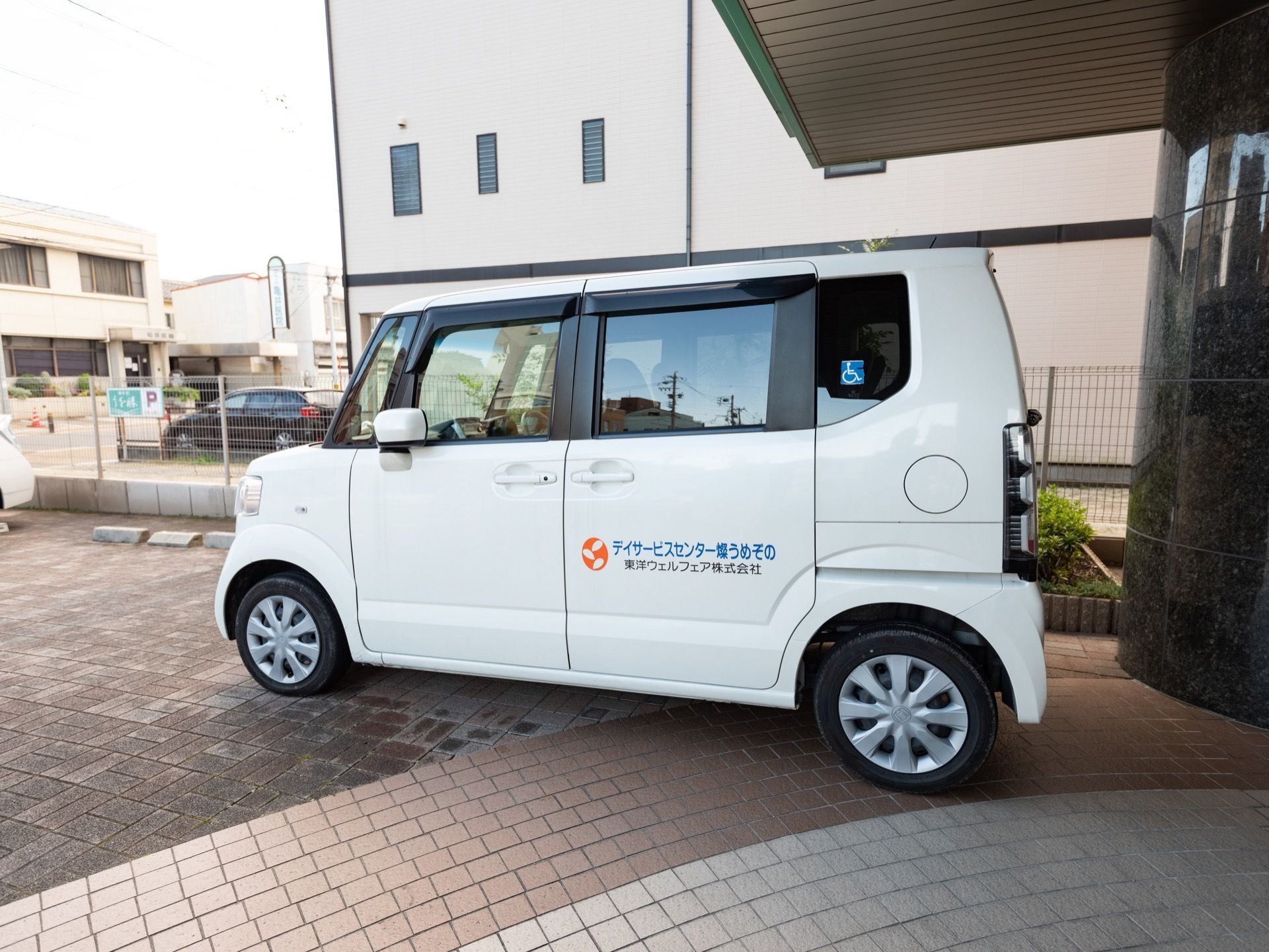 ホームの玄関前には、駐車場が設けられている。デイサービスセンターの名称などが車体に書かれた白い送迎車が１台停められている。