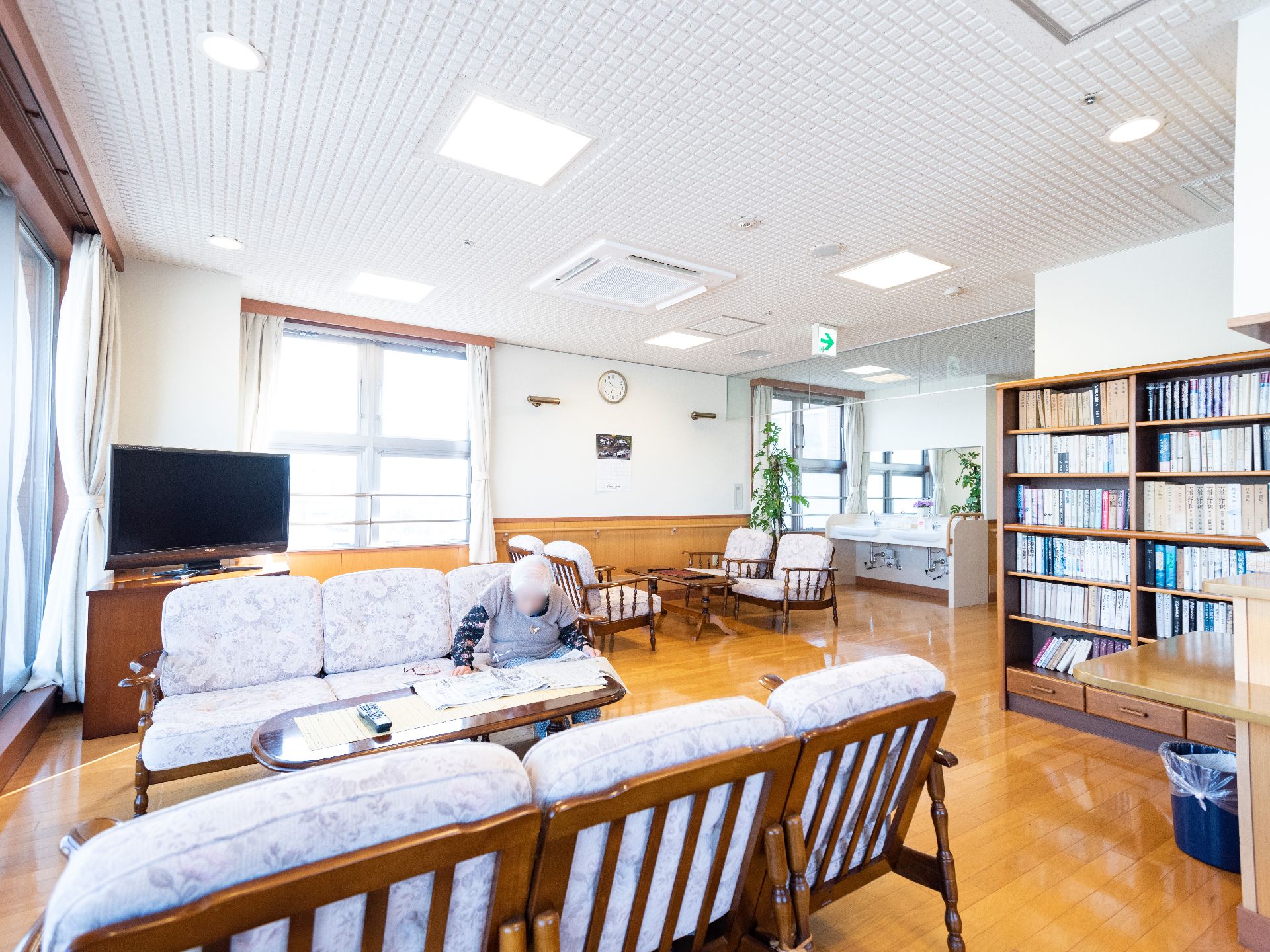 入居者様がソファに座り新聞を楽しんでいる、本棚やテレビが置かれた共有スペース