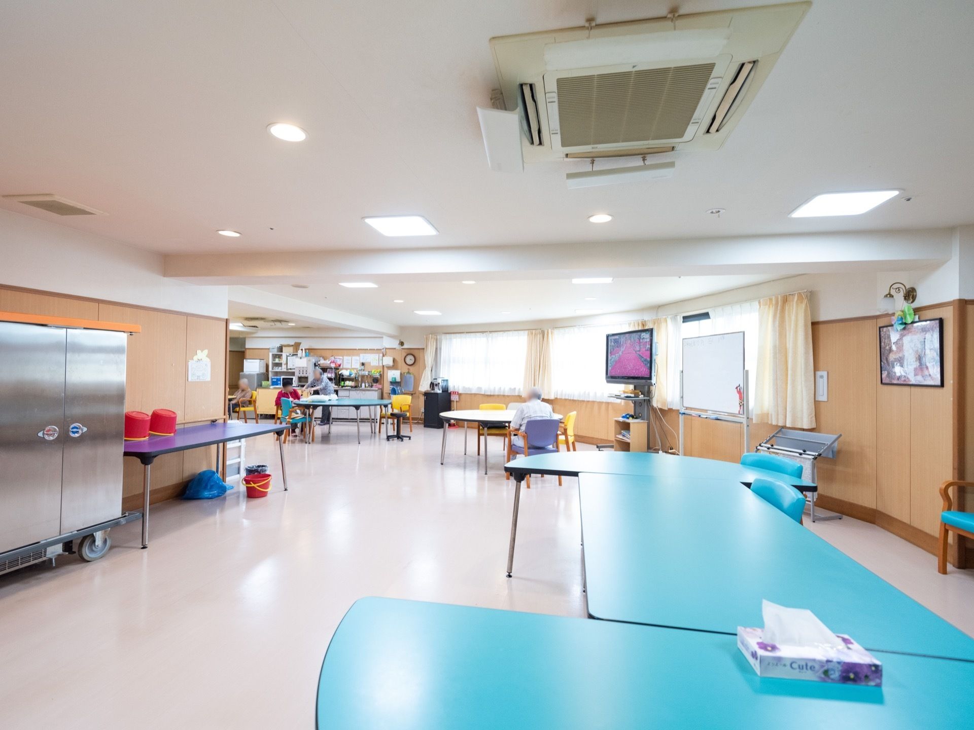 丸みを帯びた形をしている食堂である。木目調の壁で、温かみのある空間である。水色のテーブルやテレビ、ホワイトボードなどが置かれている。