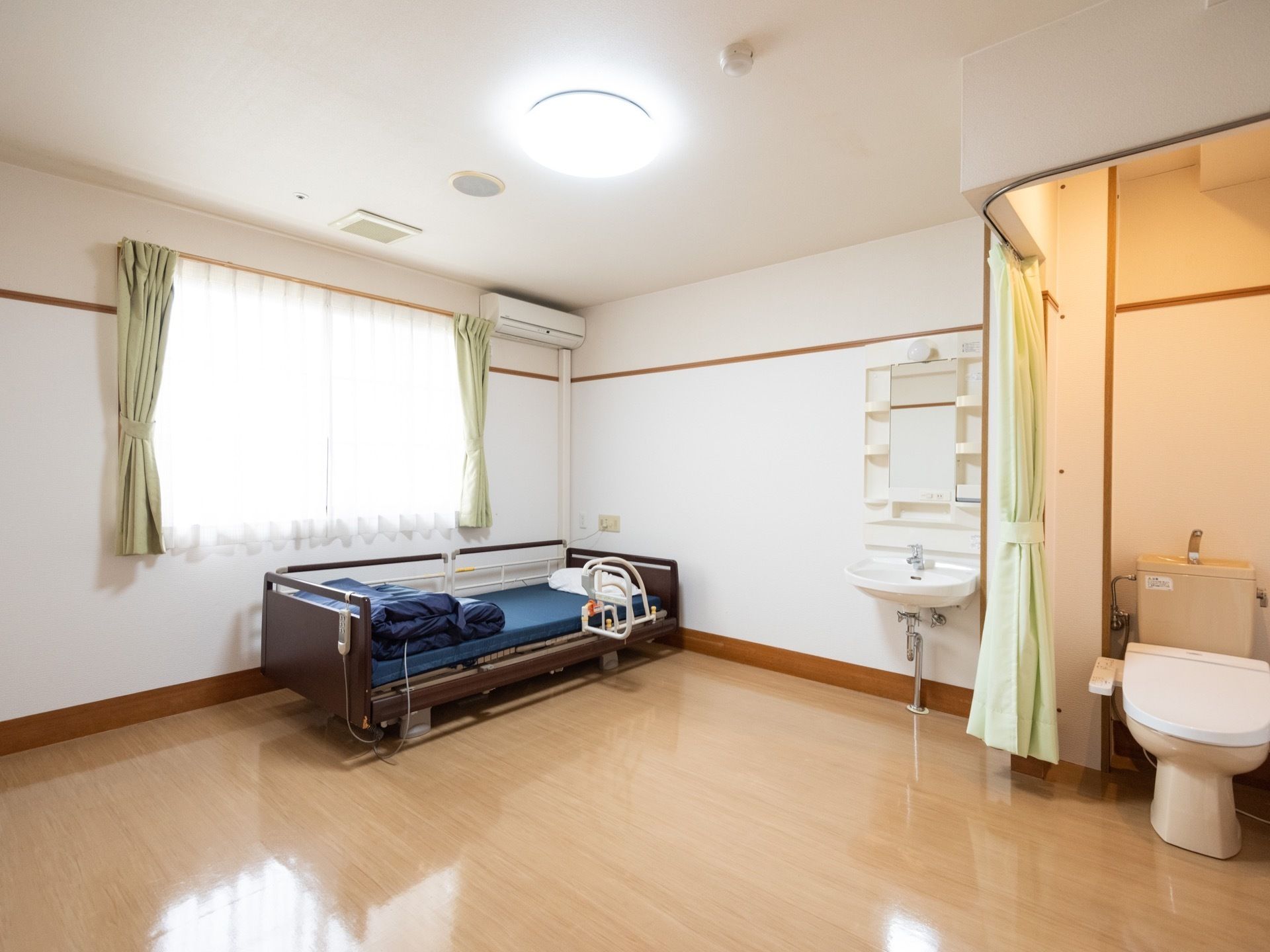 窓から差し込む光が心地よい居室である。トイレと洗面台、エアコンを設置。また、介護用のベッドも置かれている。