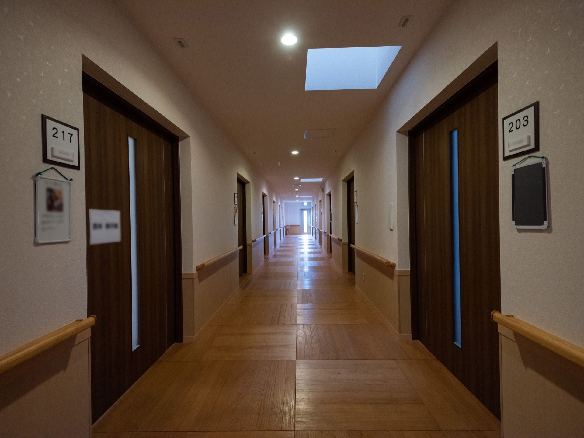 廊下はフローリングの床で、天井から壁はベージュ、手すりを境に下がピンクがかっている。明かり取りの窓がある。居室の引き戸が並んでいる。