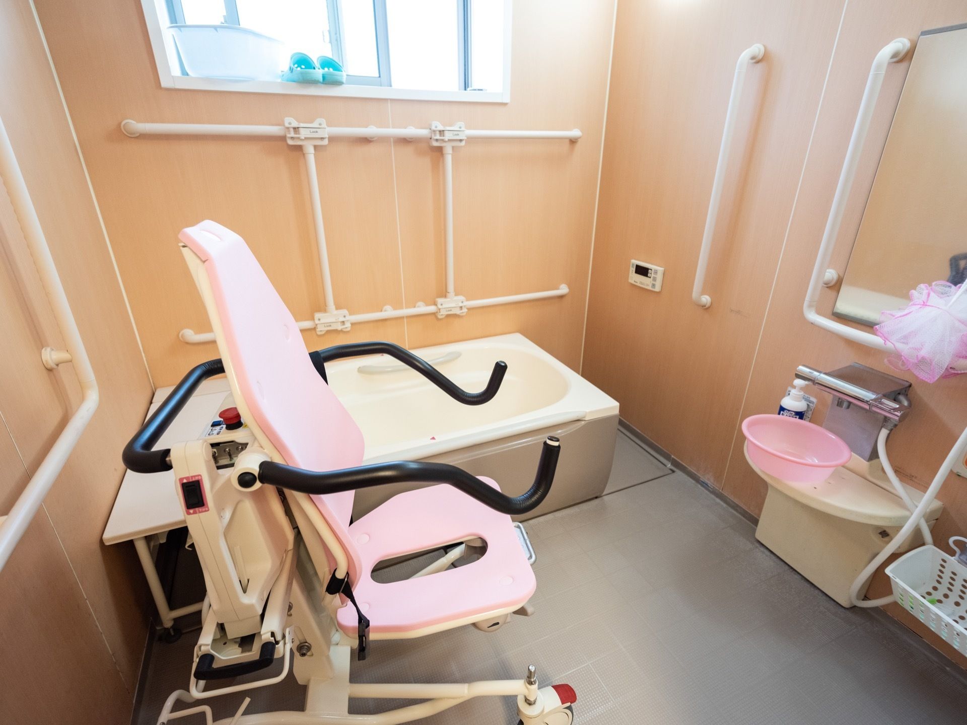 木目調の壁にグレーの床になっている浴室。入浴補助用のピンクの椅子型の搬送機がある。３方の壁面に手すりが設けられている。