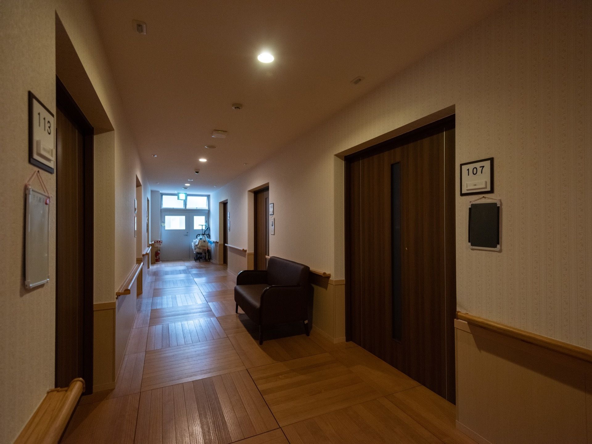 居室の引き戸が並ぶ通路が伸びていて、奥には非常口にもなっている出入り口が見える。壁面には手すりが設けられ、黒いソファが置かれている。