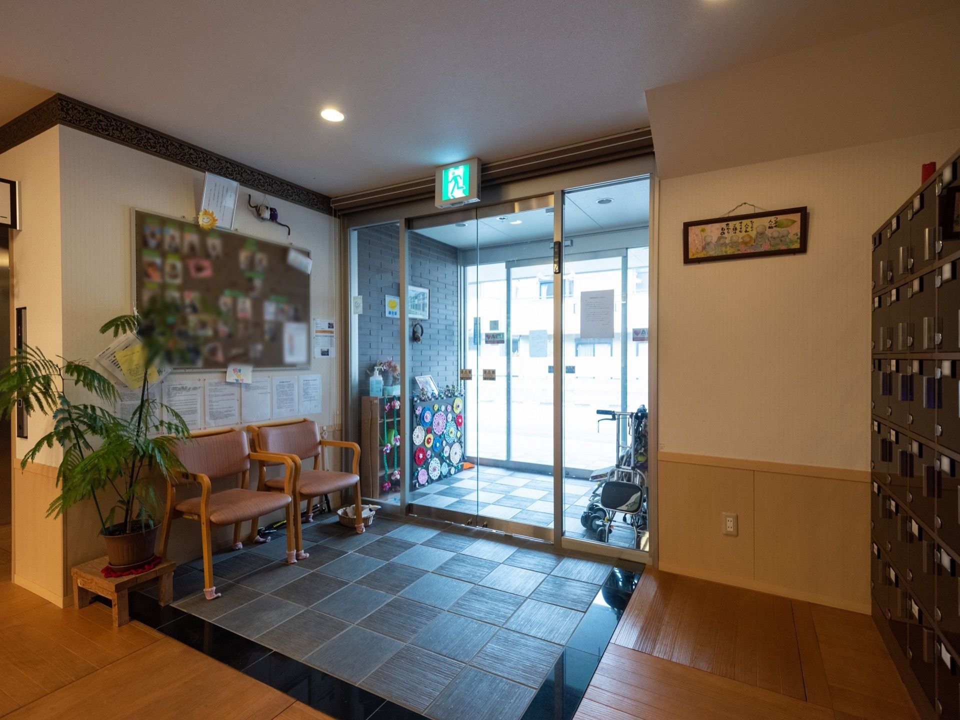二重の自動ドアが設けられている。右側の壁面に靴箱がある。三和土の部分に椅子が２つ置かれている。観葉植物を飾った置台がある。