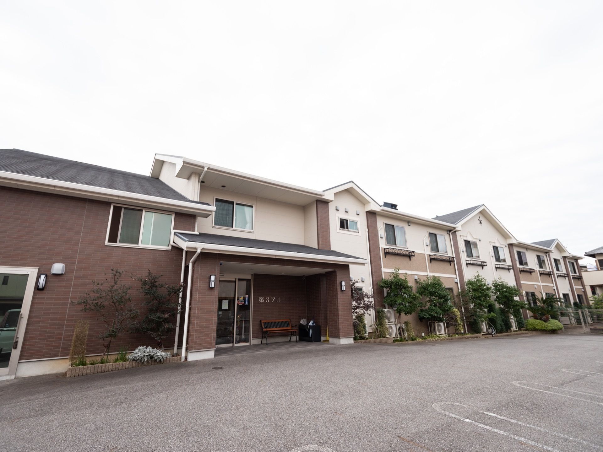 切妻屋根が組み合わされた２階建ての建物。壁は白とブラウン、ライトブラウンの３色で塗り分けられている。玄関前に舗装された駐車スペースがある。