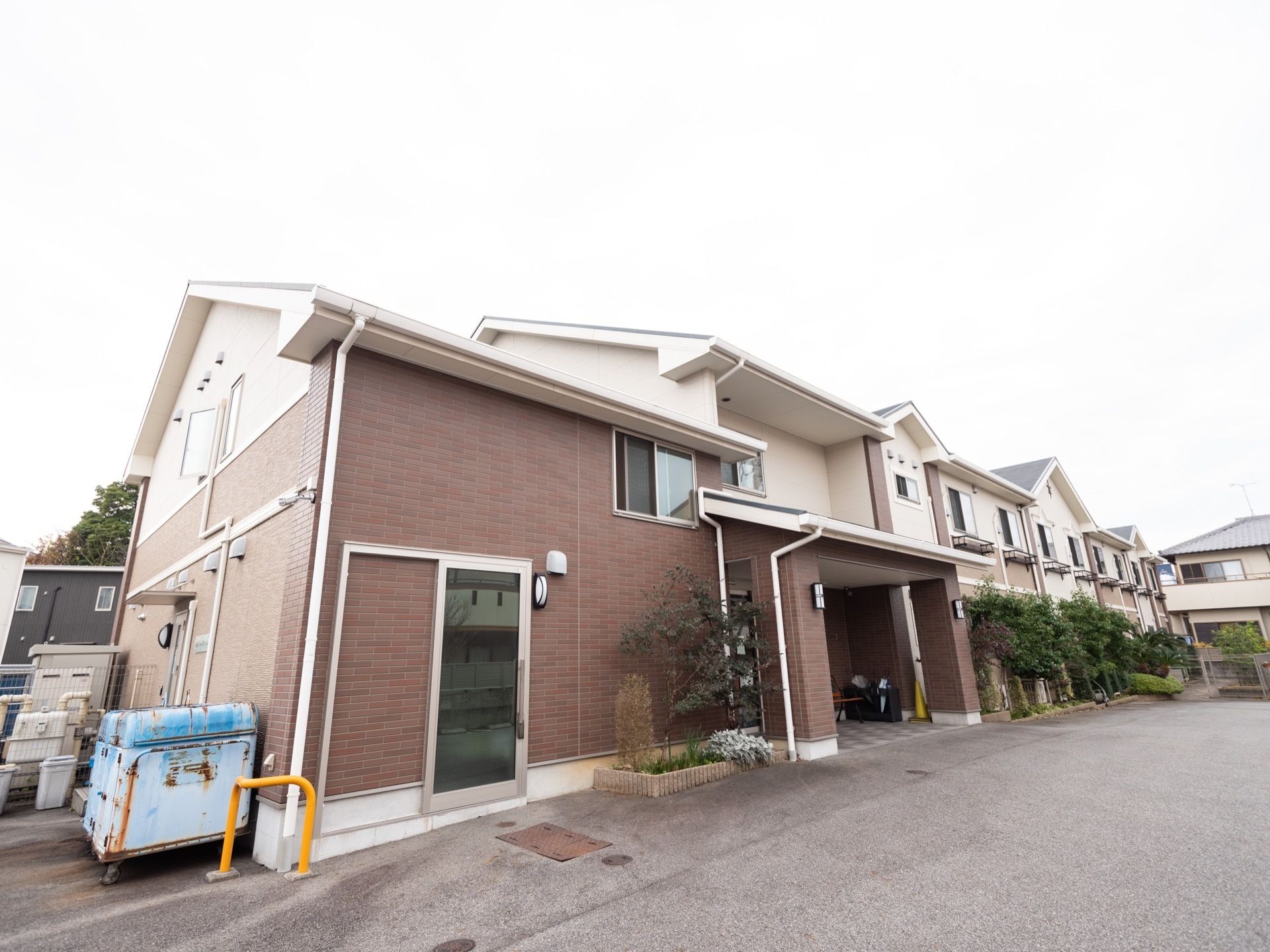 切妻屋根が組み合わさった複雑な形の２階建ての建物。玄関には大きく屋根が張り出している。通用口があり、大型のゴミ箱が置かれている。