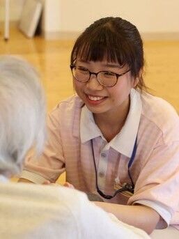 眼鏡をかけた若い女性スタッフが、笑顔で入居者に接している様子。目の高さを合わせている。