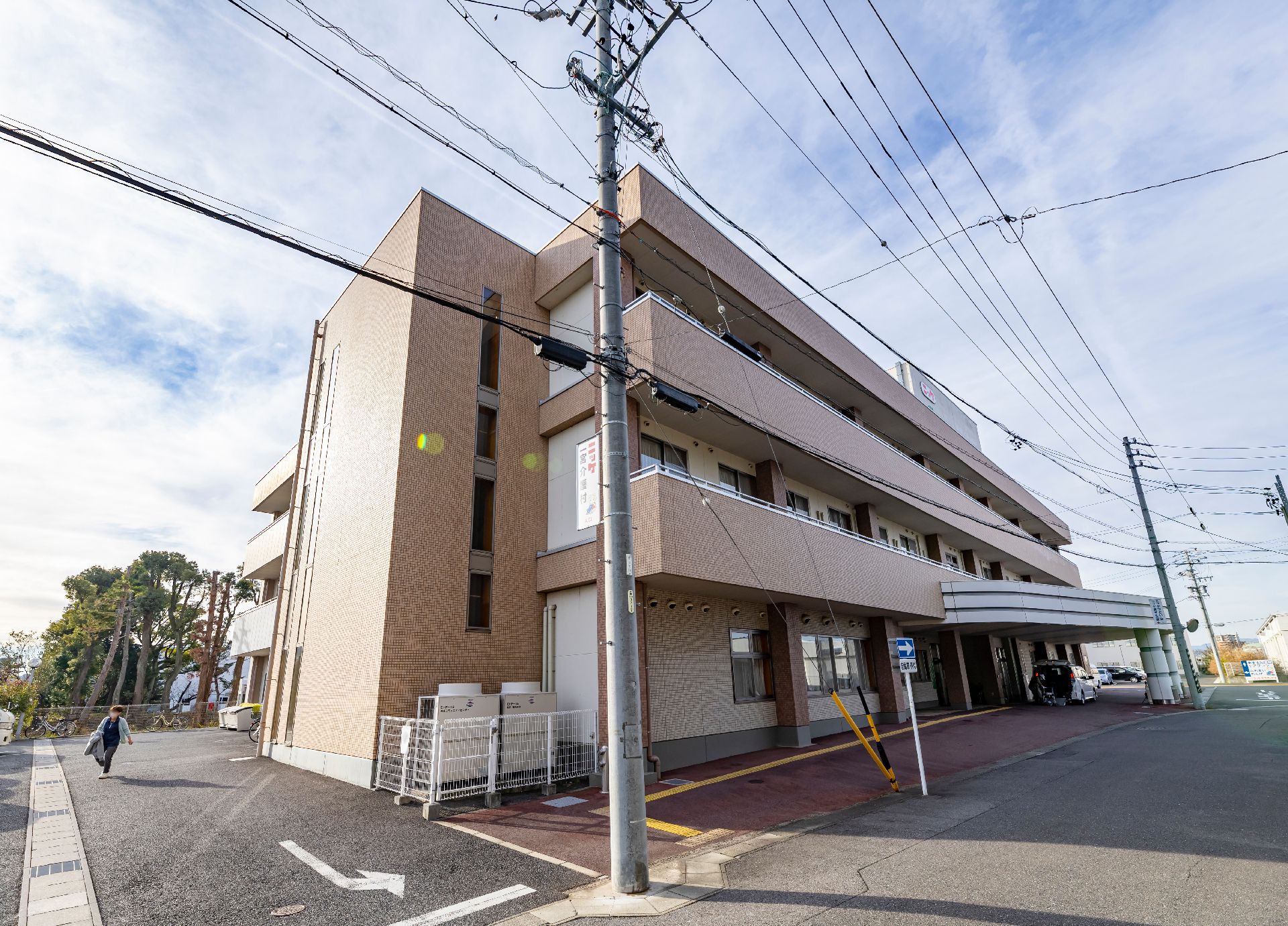 駐車場やベランダが付いたベージュの３階建ての建物。窓がたくさんつき、１階部分の出入り口には屋根が付いている。