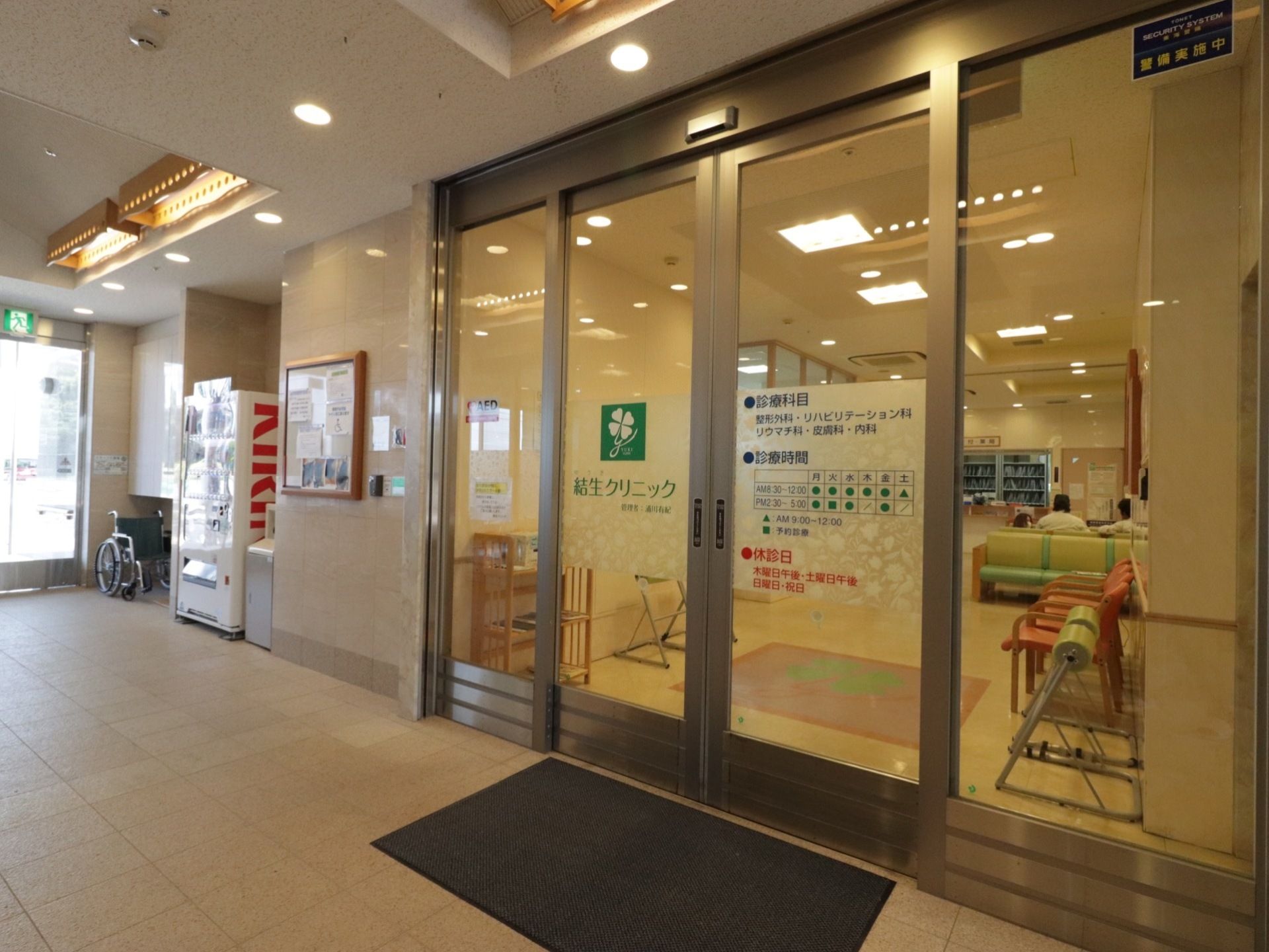 クリニック入口前の床にグレーのマットが敷かれている。横のフロアには、自動販売機や車椅子が置かれている。