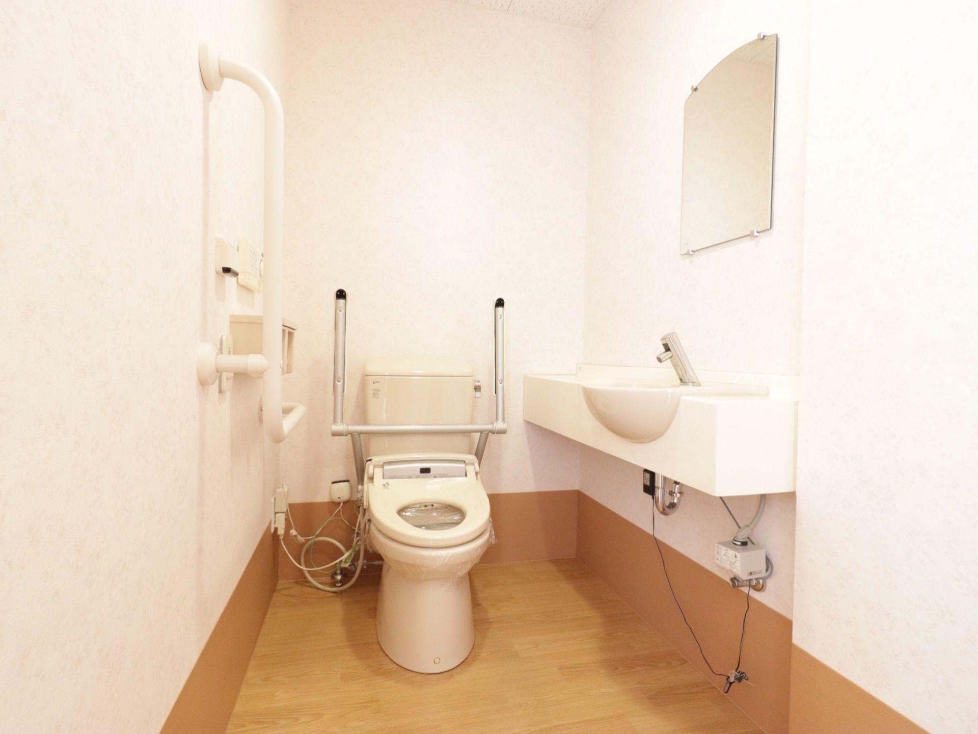 白を基調とした洗面台が設置された個室トイレである。洋式トイレの両サイドに可動式の手すりが設置されている。