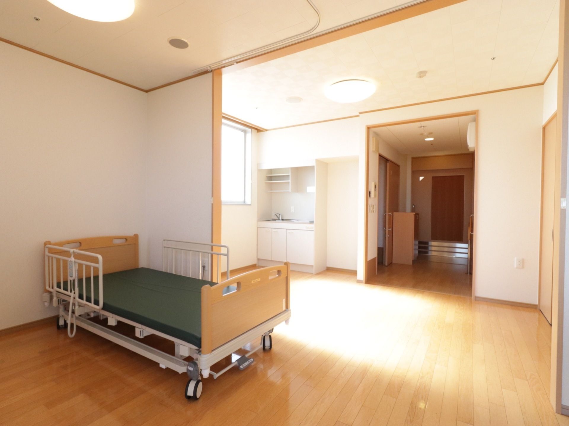 ミニキッチンが備え付けられた広々とした居室である。室内にはキャスターとリモコンが付いたベッドが置かれている。