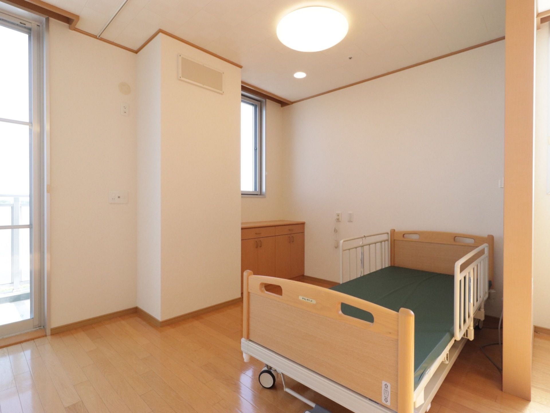 入居前の居室には、木目調の介護ベッドが設置されている。窓は大小２つあり、造り付けのチェストも配備されている。