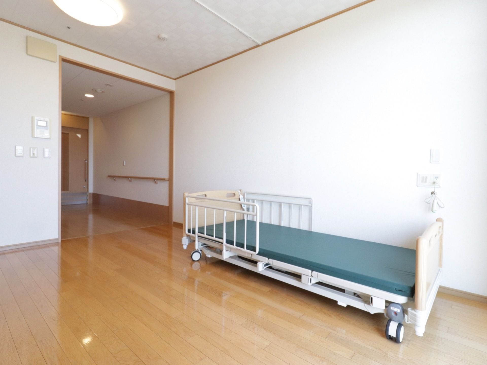 日当たりの良い居室で天井に照明が取り付けられている。壁沿いには、キャスター付きの介護ベッドが置かれている。