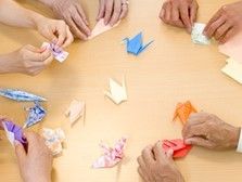 複数の人が折り鶴を折っている手のアップ。オレンジ・青・赤とカラフルな折り鶴がテーブルに置かれている。