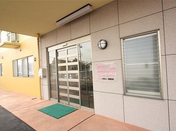 玄関のドアは屋根の下にある。ガラスの窓に、横向きの格子が入った自動ドアである。ドア上と横に照明器具がついている。