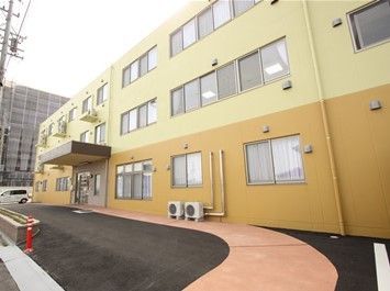 外壁が黄色の濃淡で塗り分けられた、３階建ての建物。アスファルトで舗装された施設前は、移動しやすい緩やかな傾斜になっている。