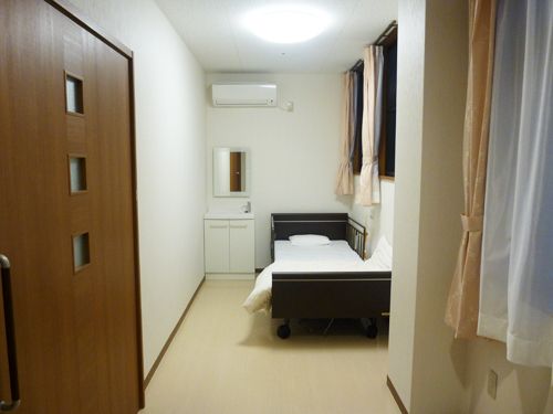 宿泊室には、介護ベッドやエアコン、洗面台が完備されている。カーテンが掛けられた窓がある。出入口の扉は引き戸になっている。
