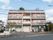 施設建物の前には、道路が通っている。外壁はピンクとホワイトの２色で、３階建てである。周囲に木が植えられている。