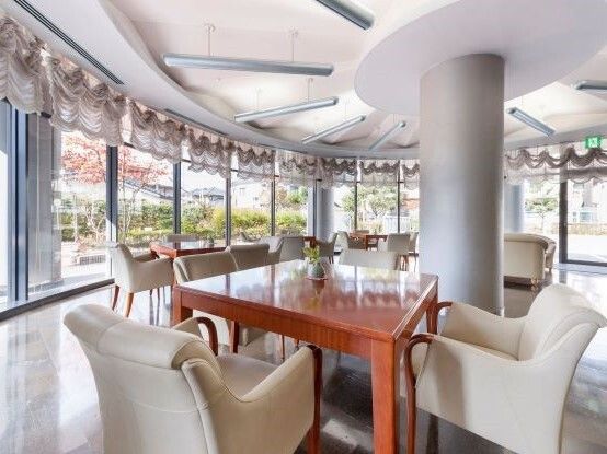 白いゆったりとした椅子がセットされた木製のテーブルが複数置かれたサロン。窓の外には中庭が見える。