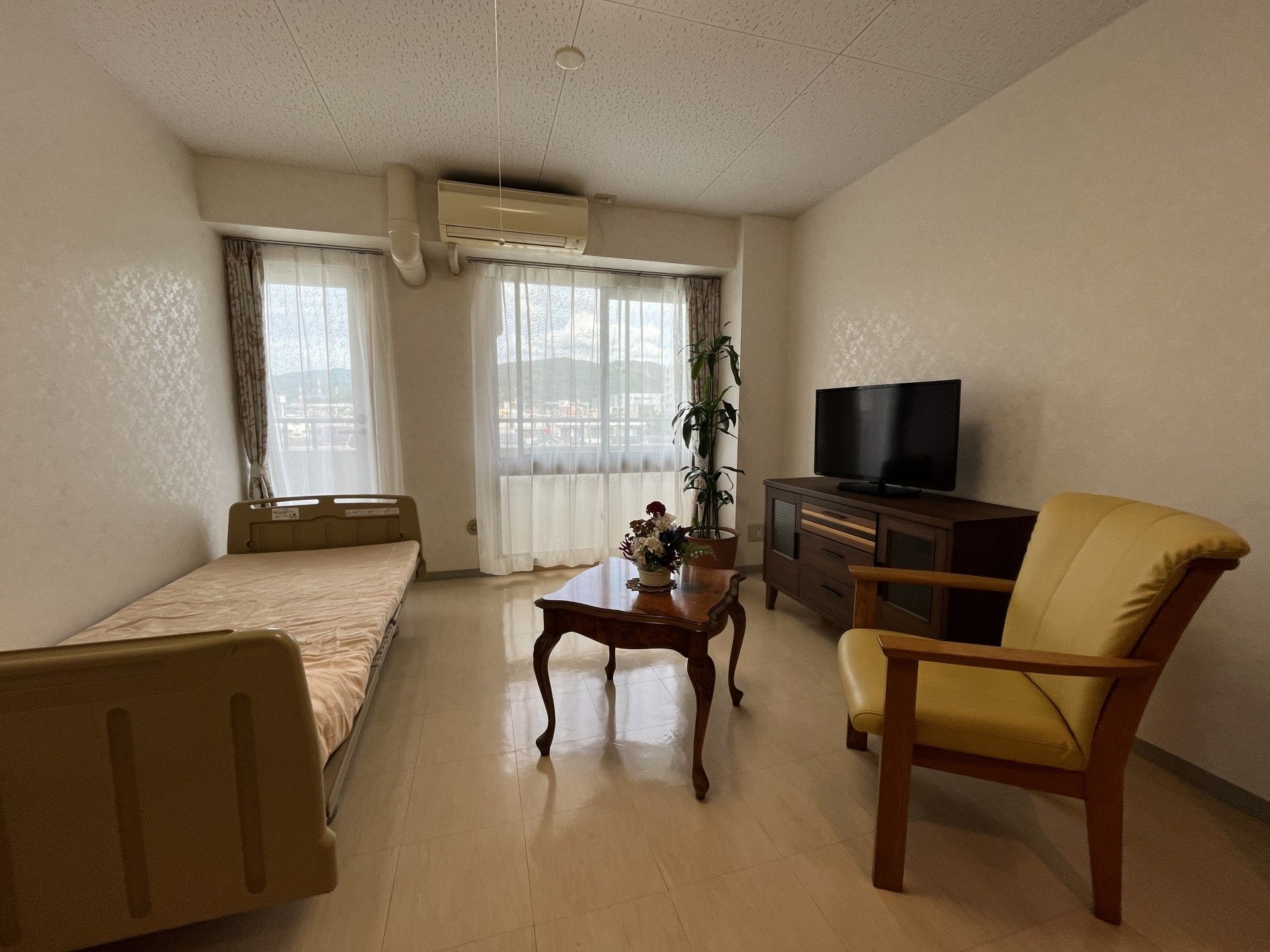 寝具付きのベッドがあり、エアコンやテレビ、一人用のいすが置かれた部屋には、カーテンが付いた窓もある。