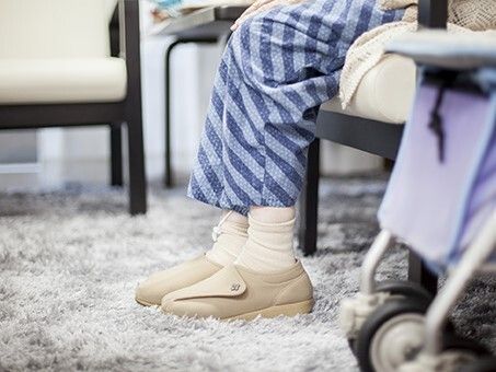 毛の長いカーペットが敷かれた部屋にはシルバーカーがあり、そばには高齢女性が座っている。足元だけの画像。