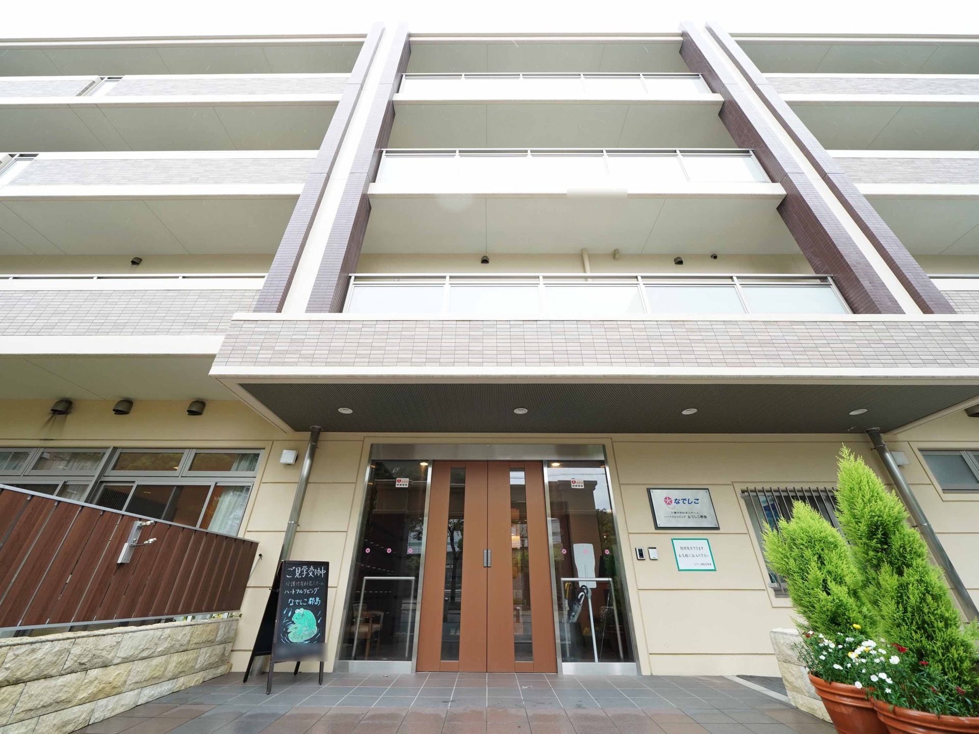 玄関は自動扉となっている。入口には黒板が置かれている。壁に施設名が表示されており、玄関前に植栽が植えられている。