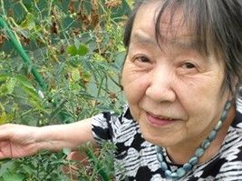 女性入居者様がゴーヤのツルに手を添えておられる。ほかにはピーマンなどの野菜が植えられたプランターもある。