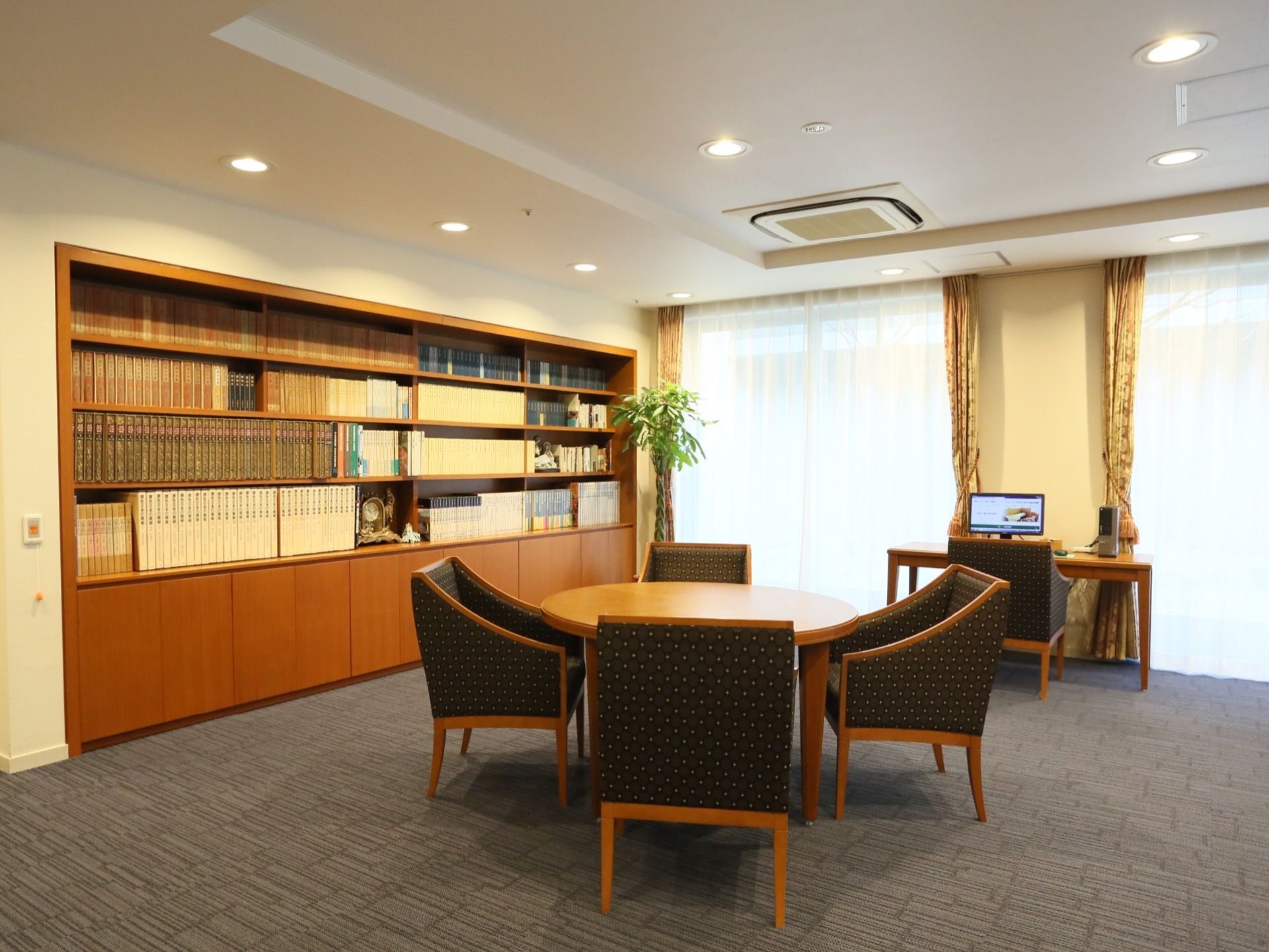 図書室には４人掛けの丸いテーブルセットと、１人用のインターネットが使用できるテーブルが備わっている。壁には大きな書棚が建てつけられている。