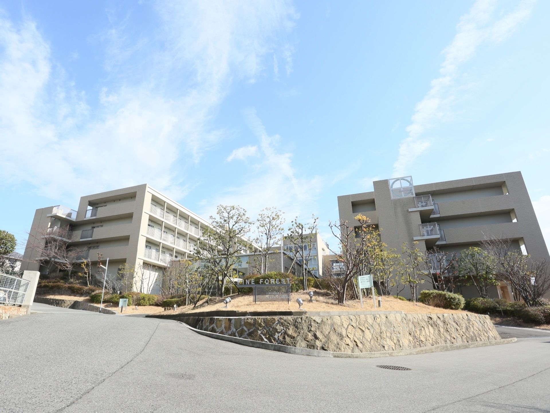 ５階建ての大きな建物が左右にあり、中心はコの字型に繋がっている。手前には樹木が植栽された大きな植え込みがあり、施設名の看板が立てられている。