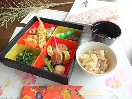 仕切りのついた弁当箱に天ぷらや焼き魚、煮物などがきれいに詰められ、炊き込みご飯と吸い物が添えられている。