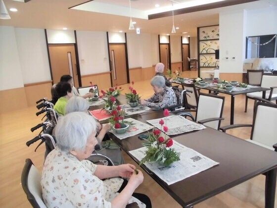 複数名の女性入居者がテーブルを囲み、花を活けている様子。車いすを利用している人もいる。
