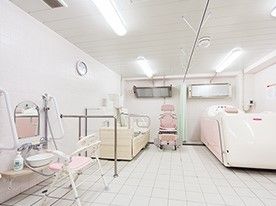 白い浴室にはピンクの機械浴槽が設置され、シャワーの前にはシャワーチェアが置かれている。
