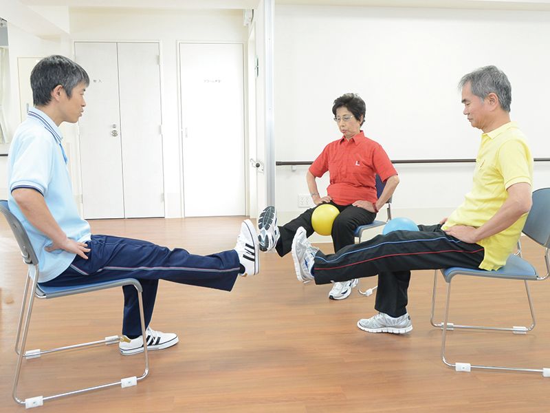 ボールを足で挟んで座り、片足を揚げている高齢の男女。前にはスタッフがいて指導している。