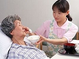ベッドにパジャマでいる高齢女性にご飯を食べさせているエプロンを付けた女性スタッフ。