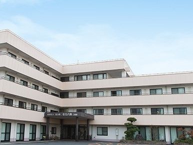 青い空の下にある白い大きな建物はエル字型で4階建て。玄関には屋根が付き、ホームの名前が書かれている。