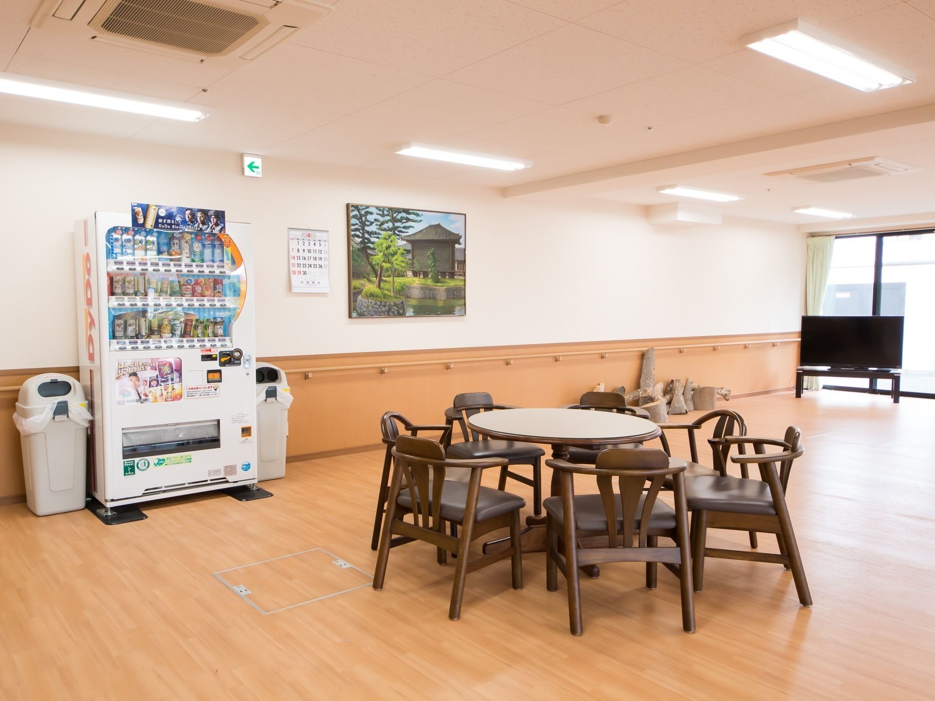 外光が差し込むホールで、自動販売機やテレビが設置されている。丸いテーブルを囲んで、椅子が複数配置されている。