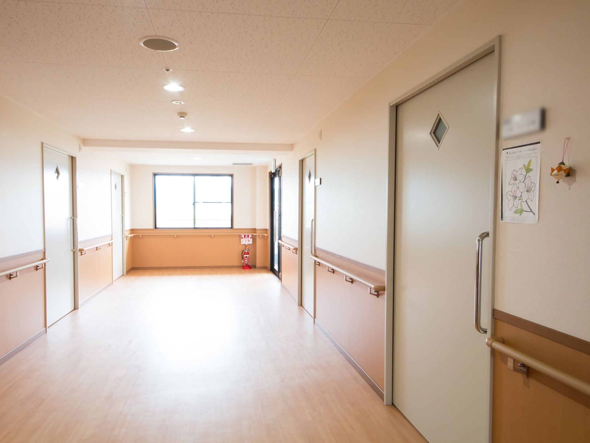 居室が立ち並ぶ広々とした廊下で、突き当たりの窓からは外光が差し込む。消火器が角に置かれており、壁には手すりが取りつけられている。