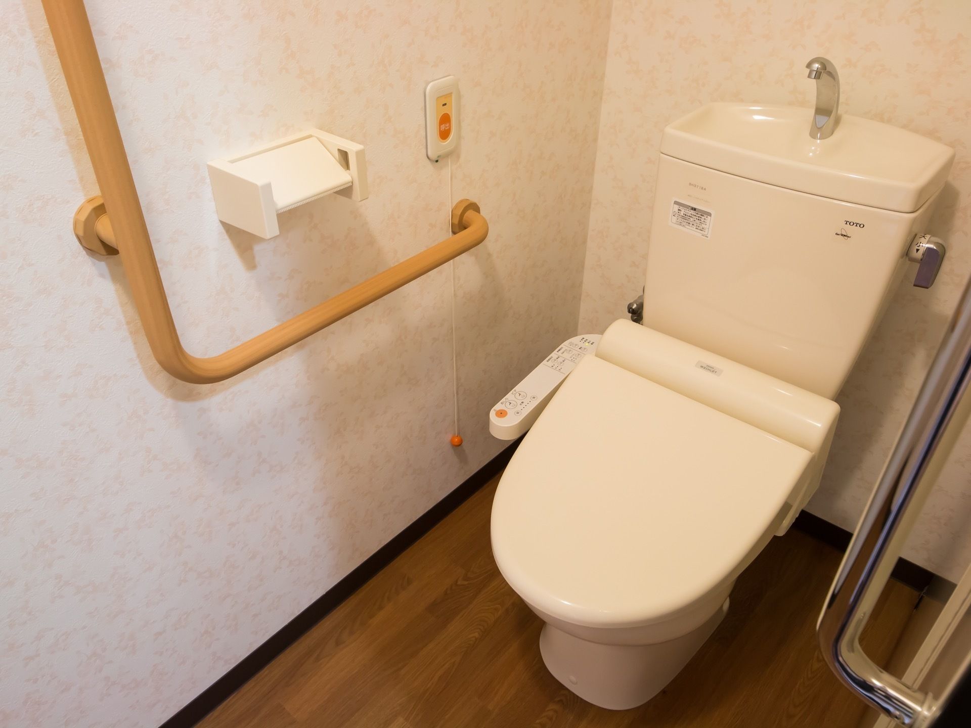 温水洗浄機能付きの個室トイレである。壁にはナースコールやＬ字型の手すり、ペーパーホルダーが設置されている。