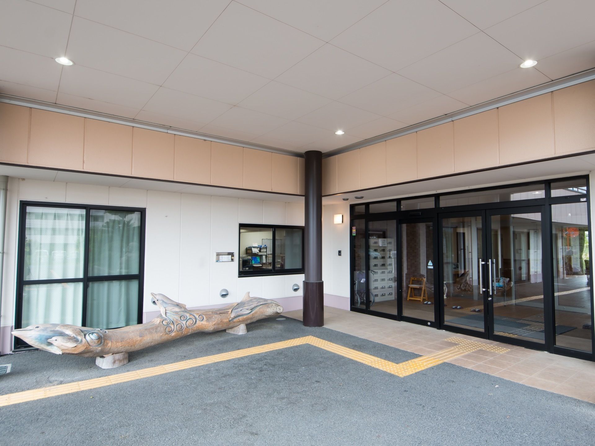 屋根付きの玄関で、地面には黄色い点字ブロックが埋め込まれている。外壁に沿って、木で作られたオブジェが置かれている。