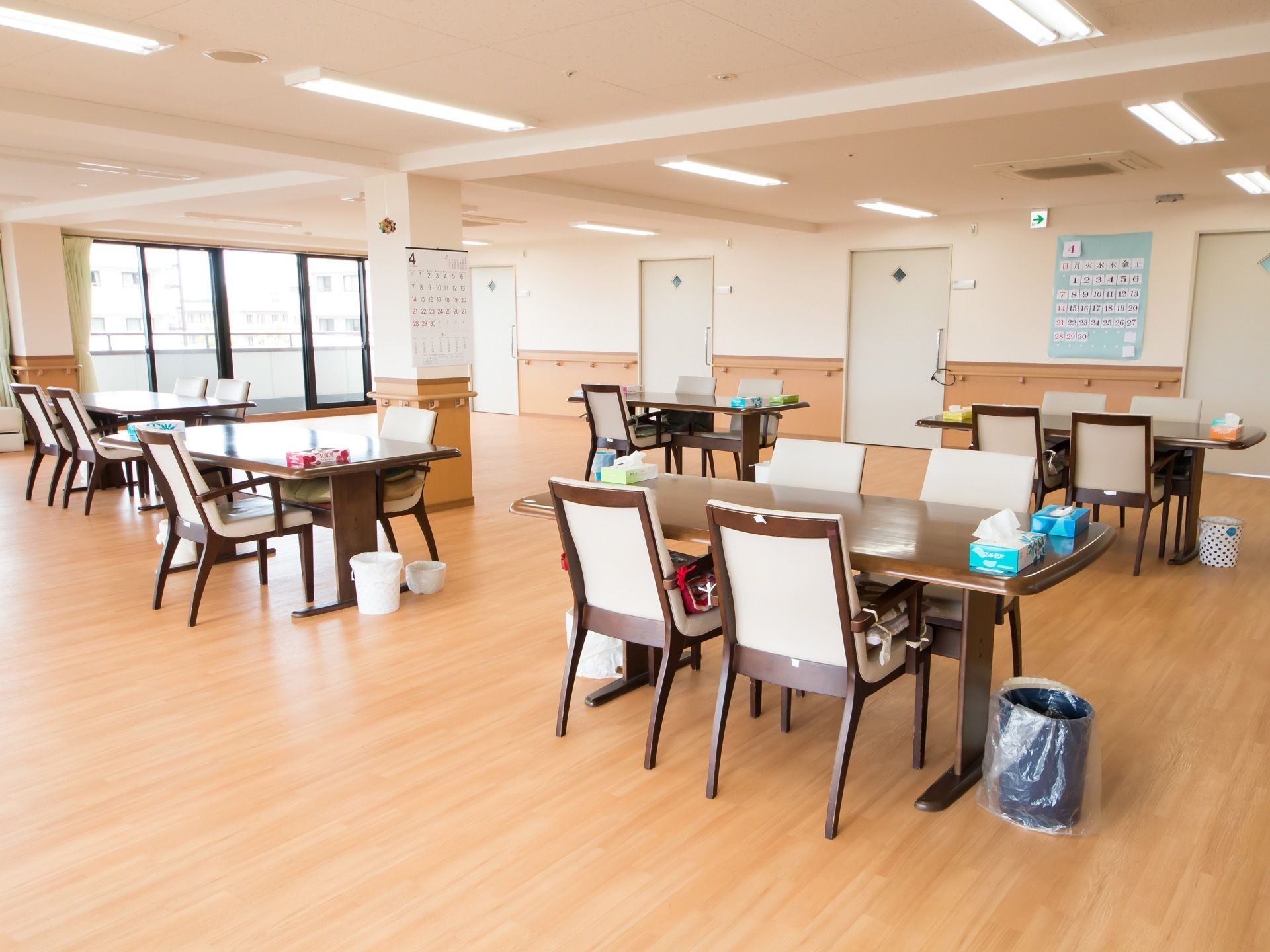 居室前に広がる開放的な食堂である。壁にはカレンダーが掛けられており、各テーブルの横にごみ箱が置かれている。